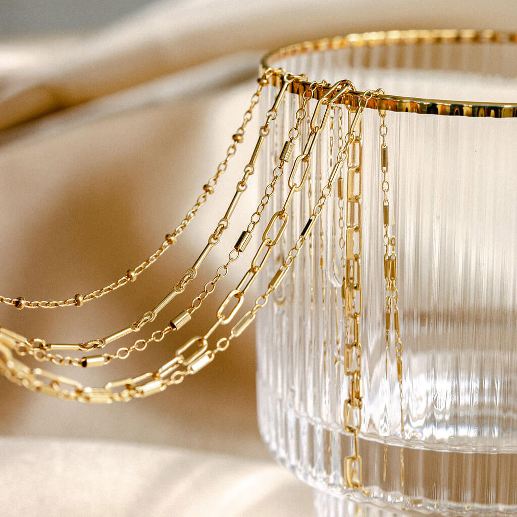 Permanent jewelry gold chains from A Royal Pampering hanging over the edge of a gold rimmed glass